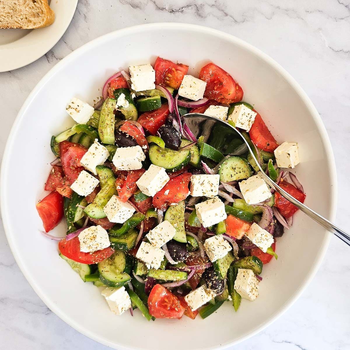 Greek salad with sliced tomatoes, cucumbers, feta cheese, olives, onions, and peppers in a white bowl