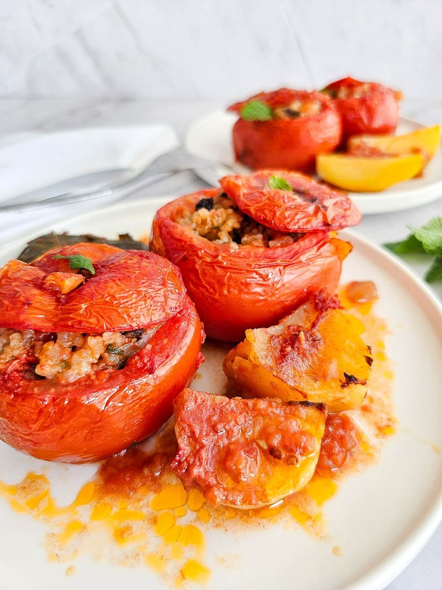 Yemista, stuffed tomatoes with rice, currants, and herbs, served with potato wedges in tomato-olive oil sauce on a white plate.